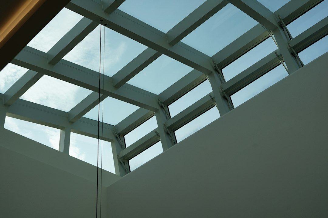 A professional installation of glass roof lanterns by Thame Windows And Doors in a contemporary home setting. In the foreground, skilled technicians in smart work uniforms are carefully lifting and positioning a large, sleek glass lantern into place, showcasing precision and teamwork. The middle ground features the partially installed lantern surrounded by scaffolding, highlighting the expertise in roof installation. The background captures a serene, cloudy sky, allowing soft, diffused light to enhance the glass's reflective quality. The atmosphere conveys professionalism, craftsmanship, and a focus on detail, with a lens angle that emphasizes the height and structure of the lantern against the home's architecture. The image is bright and inviting, showcasing quality work in a residential environment.