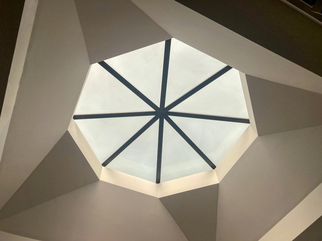 A beautifully designed glass roof lantern, featuring an elegant geometric structure made of clear glass and brushed aluminum framing. The lantern is positioned prominently in the foreground, showcasing its intricate details and modern aesthetics. Light pours through the transparent panels, casting soft reflections and illuminating the surrounding area. In the middle ground, a charming view of a cozy living space is visible, with stylish furniture and lush greenery peeking through large windows. The background features a serene sky, hinting at dawn or dusk with soft pastel hues. The mood is peaceful and inviting, highlighting the benefits of natural light and open spaces. Inspired by Thame Windows And Doors, focus on its elegance and functional design for enhancing interiors.